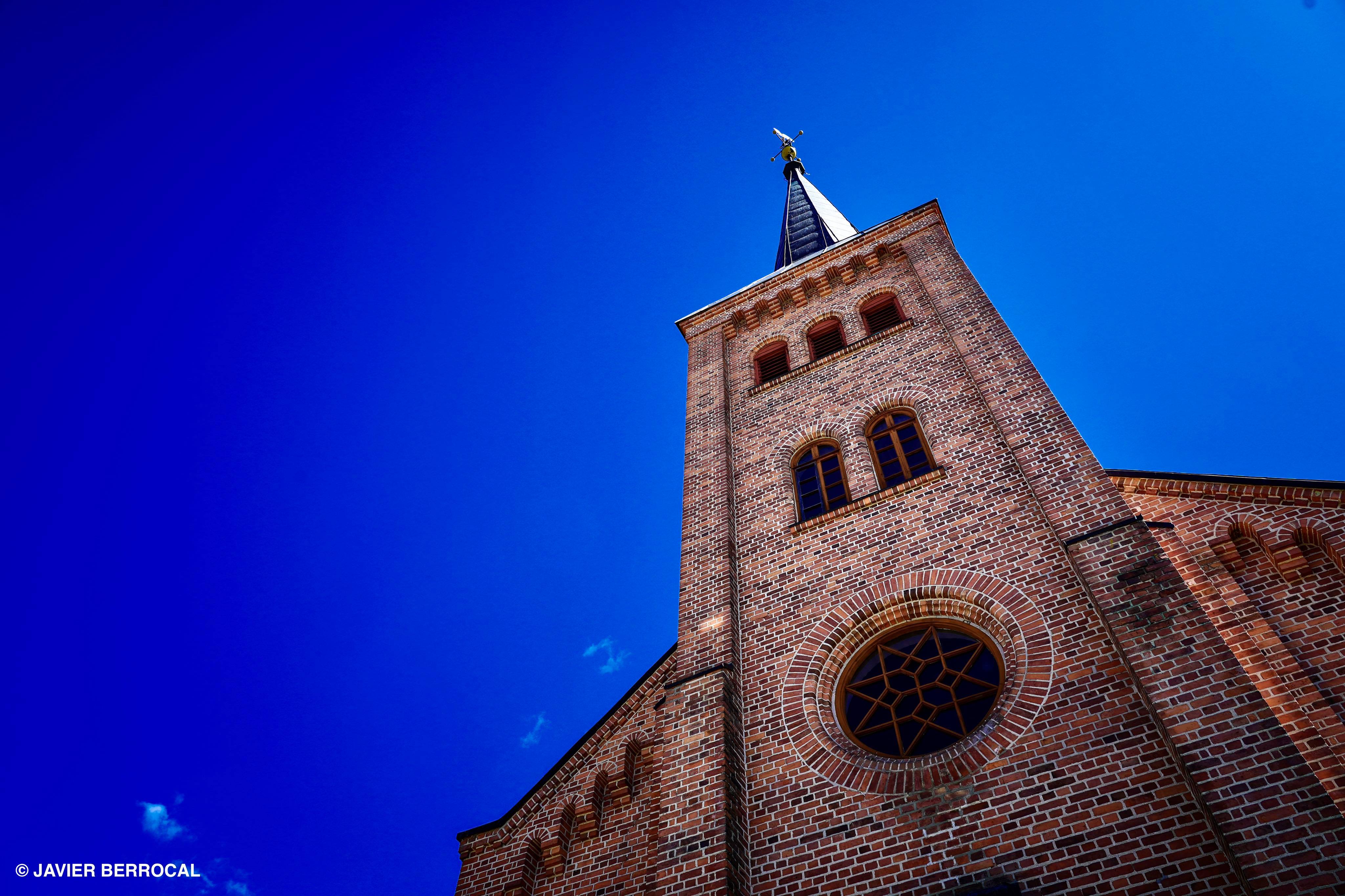Nes kirkes fasade, sett nedenfra og litt på skrå, med klokketårn ruvende mot knallblå himmel