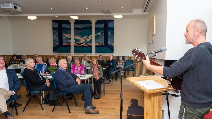 Mann med gitar synger og spiller for voksne mennesker som sitter ved flere bord rundt i lokalet